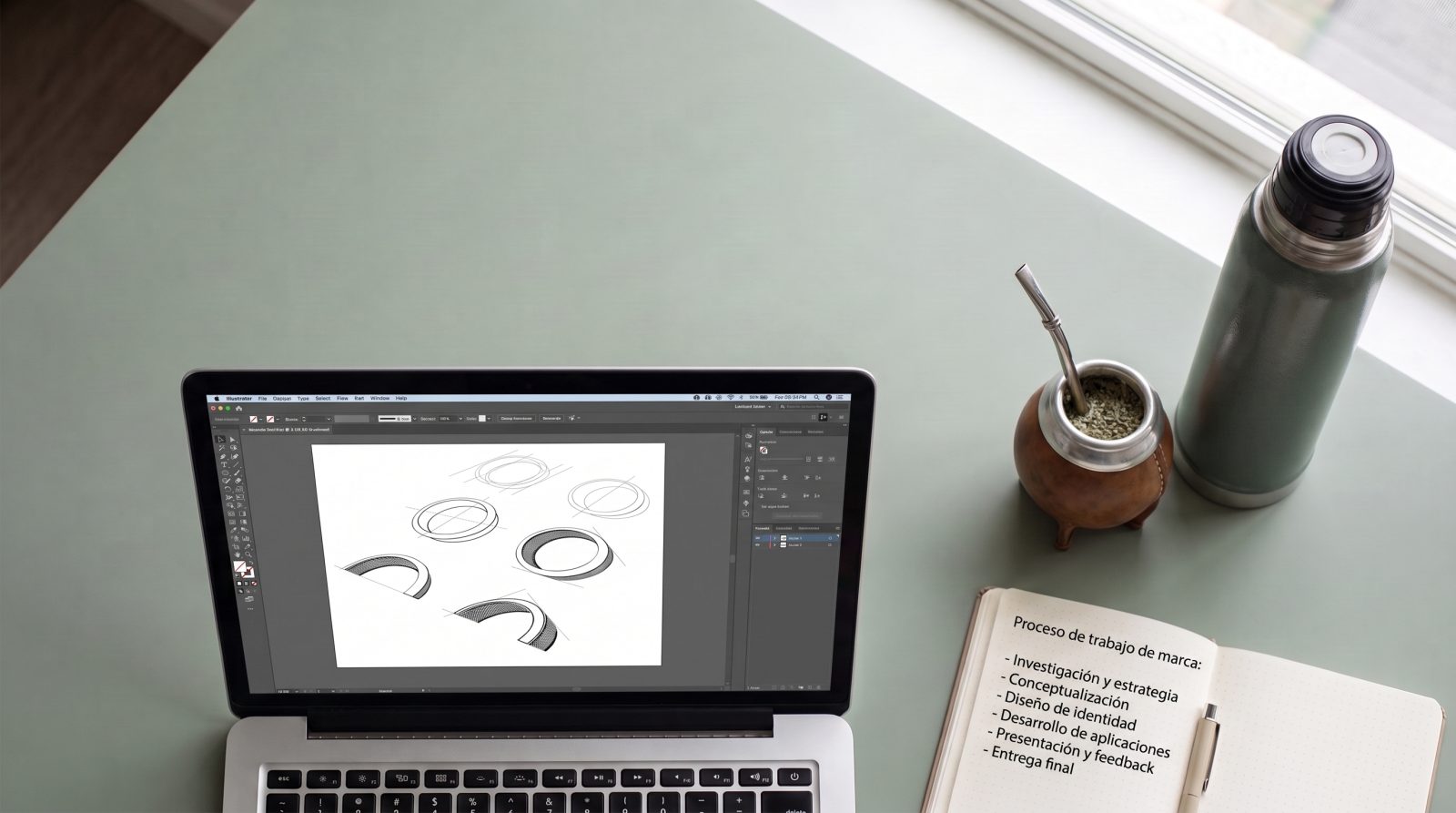 Escritorio del estudio: bocetos de un logo en Illustrator en la notebook, un cuaderno con el proceso de trabajo de marca anotado a mano y un mate.